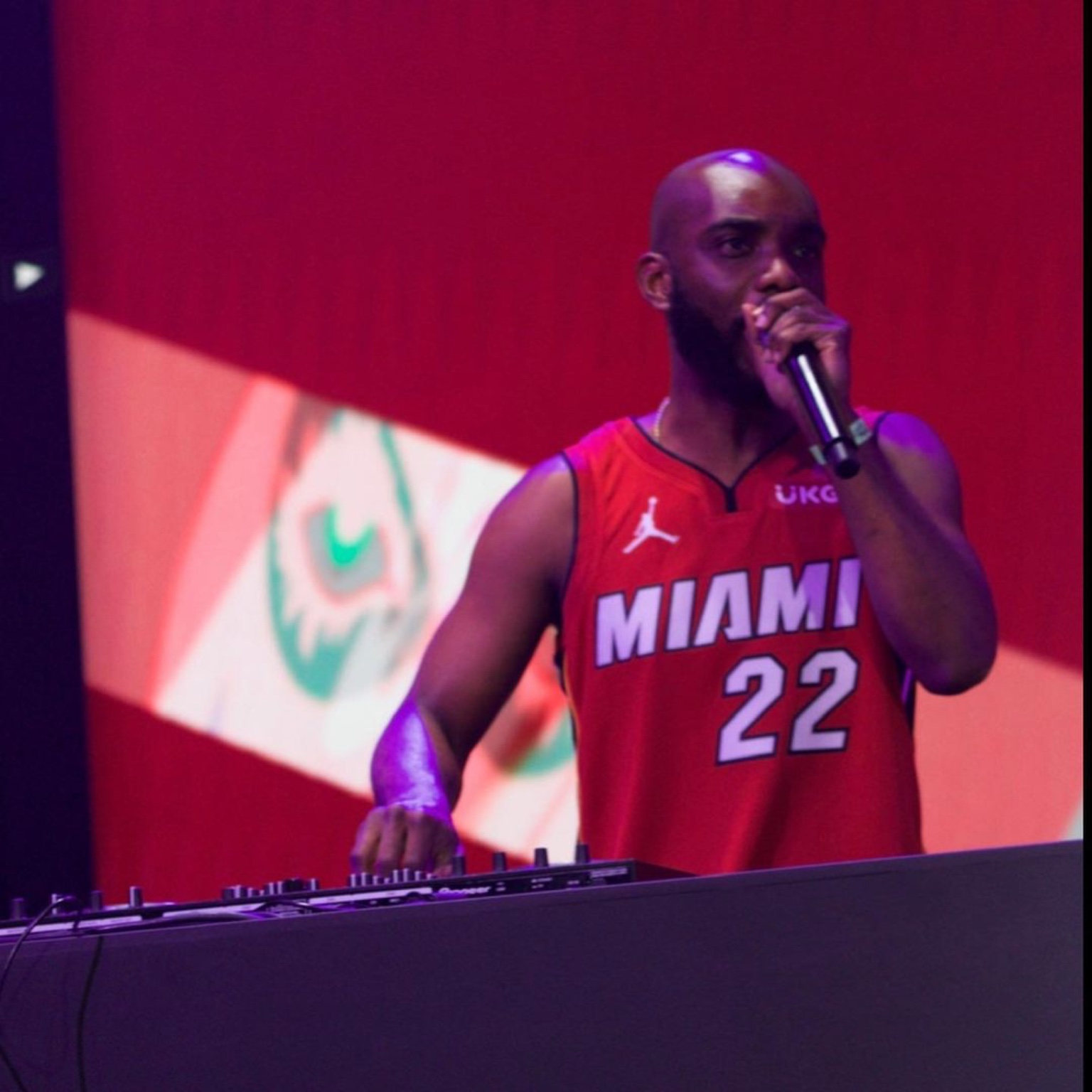 Radio Jamez DJing and holding a microphone on stage in a red Miami jersey